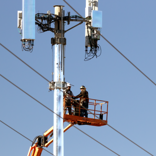 Technicians on a lift installing communication devices on a utility pole against a clear sky.